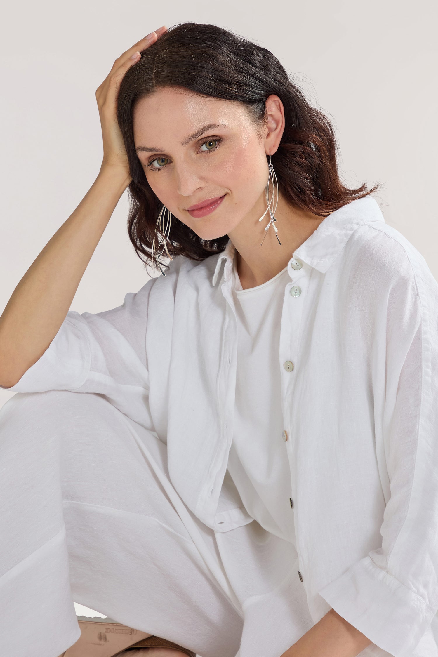 A person with shoulder-length dark hair, wearing a white Etnika outfit and large Arc Silver Earrings, seated and resting their head on their hand, smiles at the camera.