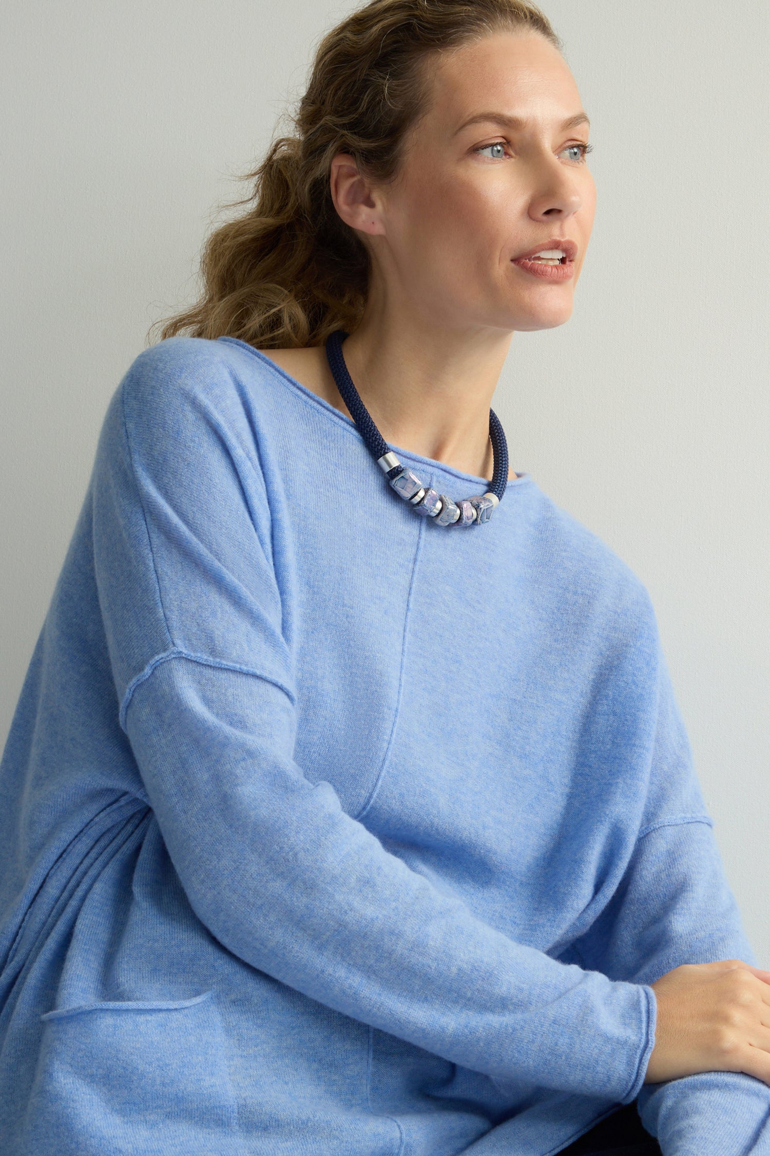 The individual, thoughtfully gazing sideways, is adorned in a light blue sweater paired with the artisanal Mosaic Cube Cord Necklace.