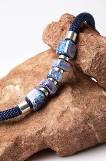 The Mosaic Cube Cord Necklace, featuring navy braided rope and silver-blue patterned beads crafted artisanally, is elegantly draped over a large brown rock.