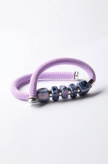 The Mosaic Cube Cord Necklace features purple braids with blue and silver beads, highlighting artisanal craftsmanship against a light backdrop.