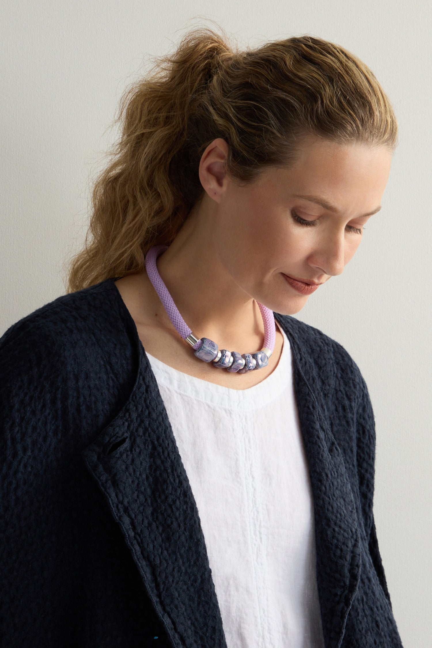 The person with wavy hair gazes down, wearing a textured black jacket over a white top. Around their neck is the Mosaic Cube Cord Necklace—a purple rope adorned with silver beads, showcasing artisanal craftsmanship.