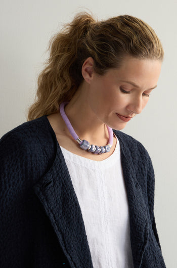 The person with wavy hair gazes down, wearing a textured black jacket over a white top. Around their neck is the Mosaic Cube Cord Necklace—a purple rope adorned with silver beads, showcasing artisanal craftsmanship.