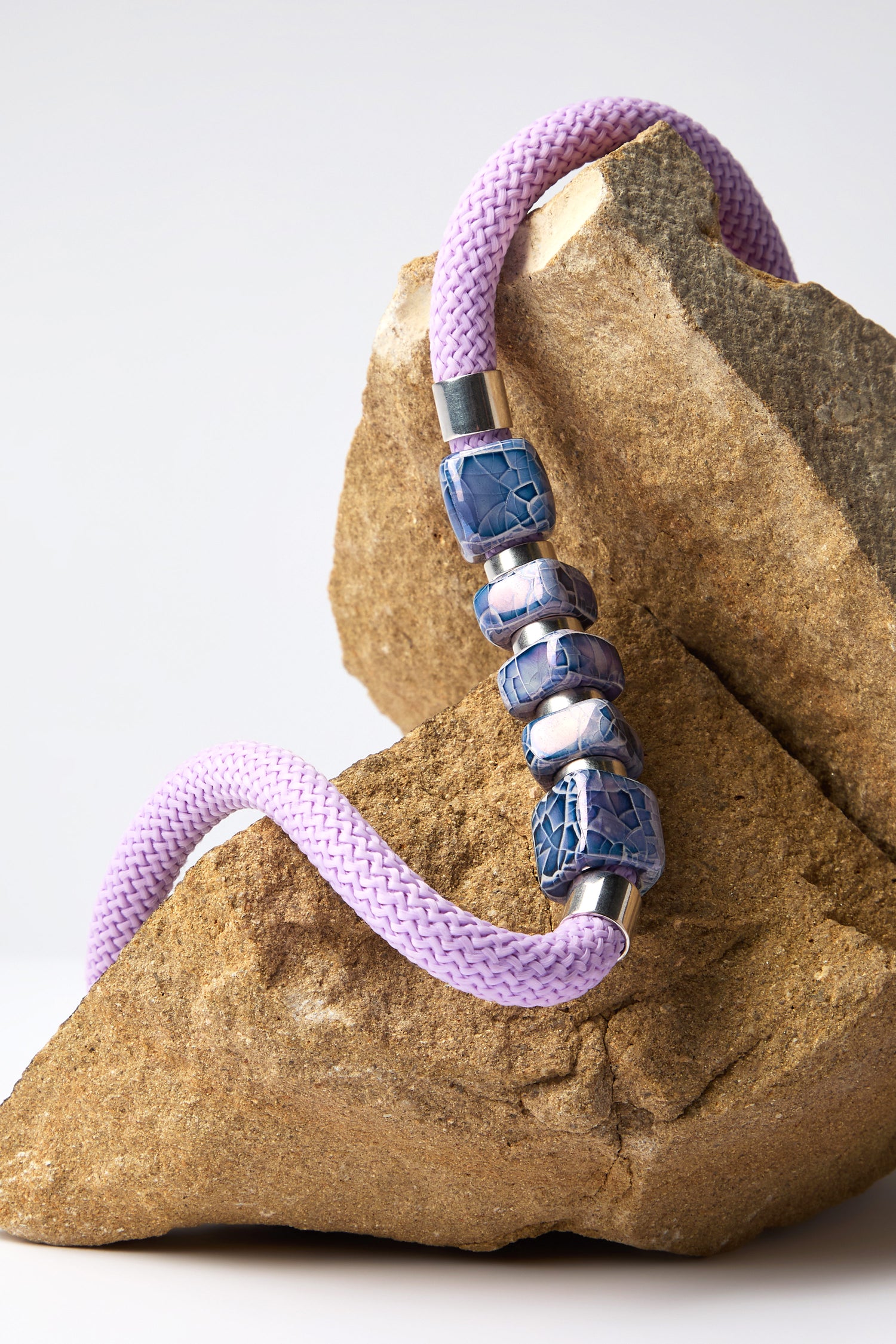 The artisanal Mosaic Cube Cord Necklace showcases a lavender braided rope adorned with blue beads, elegantly draped over a large, rough brown rock, creating a stunning statement piece.