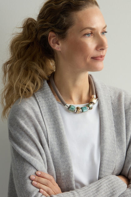 A woman with light brown hair in a ponytail, wearing a gray cardigan and white shirt, glances sideways. Her style is elevated by the Marble Cube Cord Necklace from Etnika, subtly transforming her attire into a contemporary statement piece.