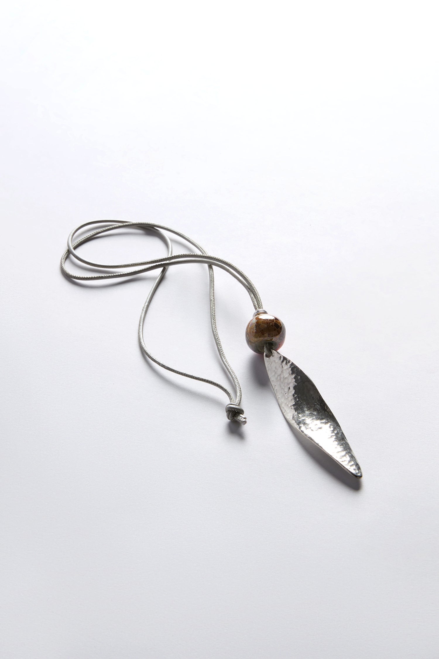 The Twisted Leaf Leather Cord Pendant rests elegantly on a light surface, showcasing a long, metallic design with a brown bead that seamlessly blends modern style with organic shapes.