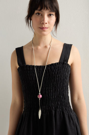 A woman wears a black textured sleeveless dress with a long Etnika Jewelry Twisted Leaf Leather Cord Pendant necklace featuring a pink bead, standing against a plain backdrop.