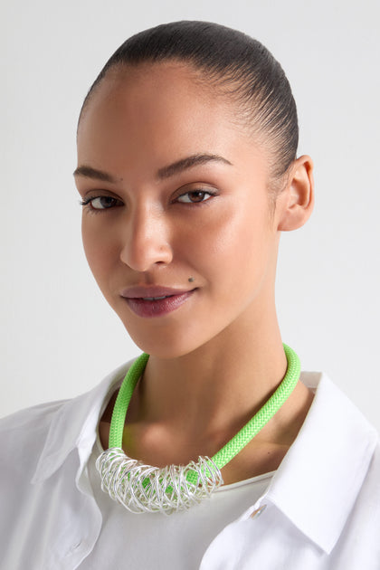 A woman with a slicked-back hairstyle is wearing the Silver Scribble Cord Necklace, featuring an abstract silver pendant on an emerald green cord, paired with a white shirt and smiling slightly.