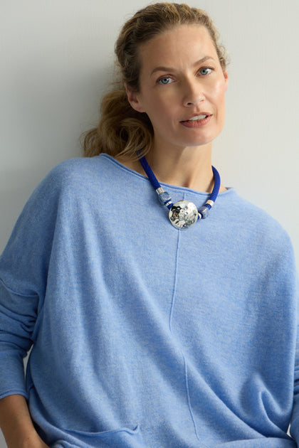 Against a plain background, a person in a blue sweater displays the artisanal Hammered Circle Cord Necklace, defined by its large blue hammered metallic circular pendant.