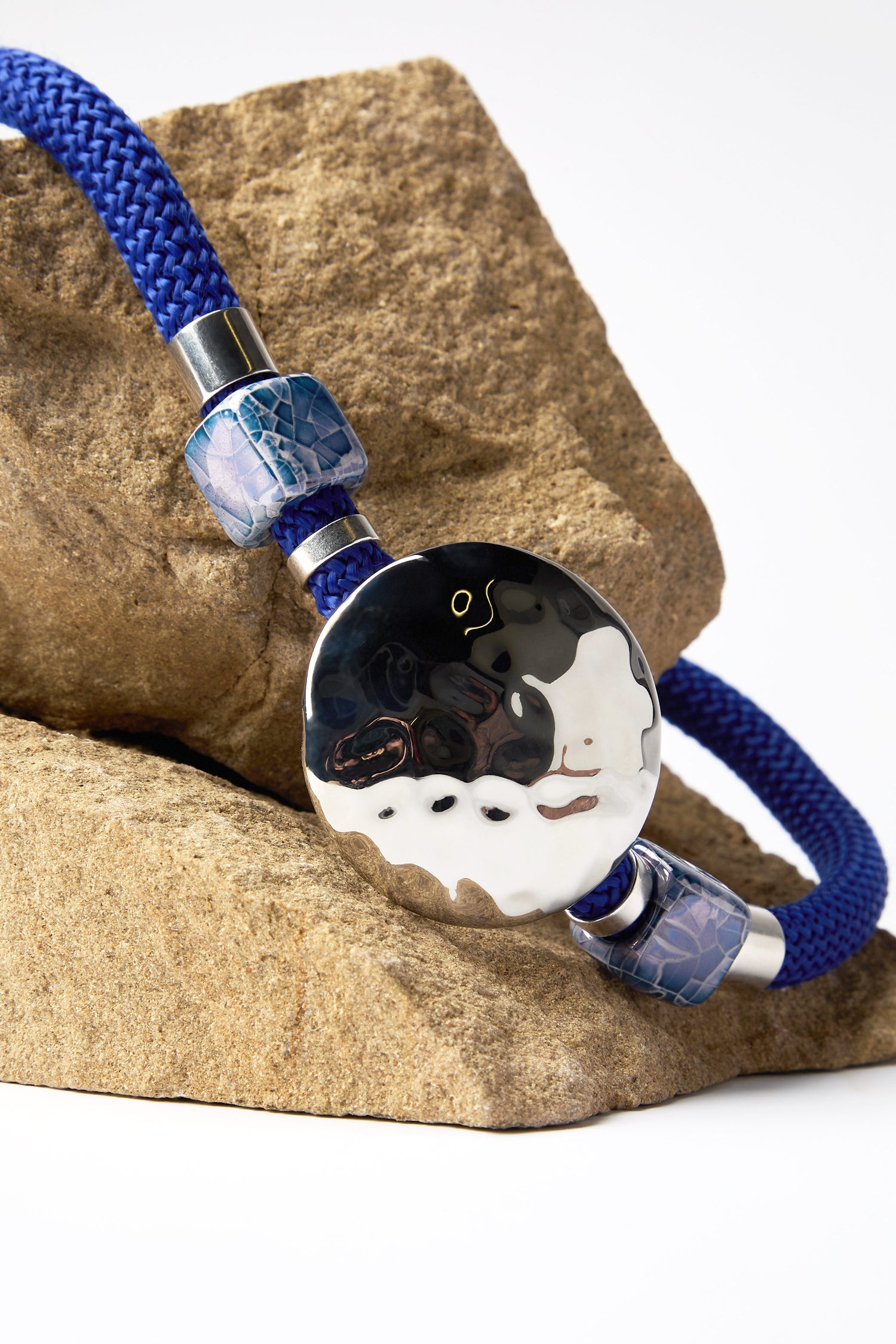 The Hammered Circle Cord Necklace showcases a round black and white ceramic bead, accented by two smaller blue decorative beads, elegantly placed on a rock.