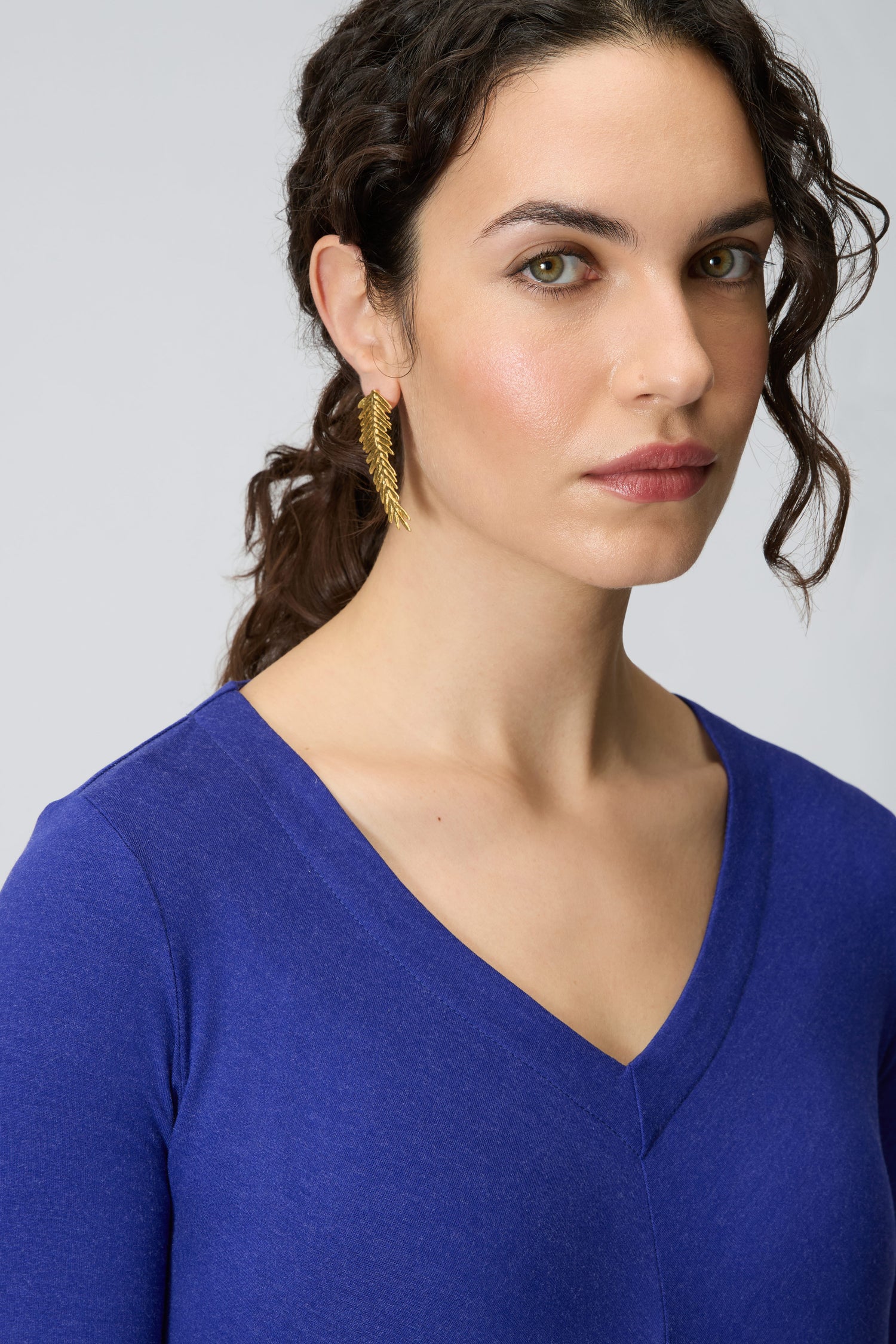 A woman with wavy dark hair in a ponytail wears a blue V-neck top and Gold Plated Feather Earrings, looking slightly past the camera with a neutral expression.