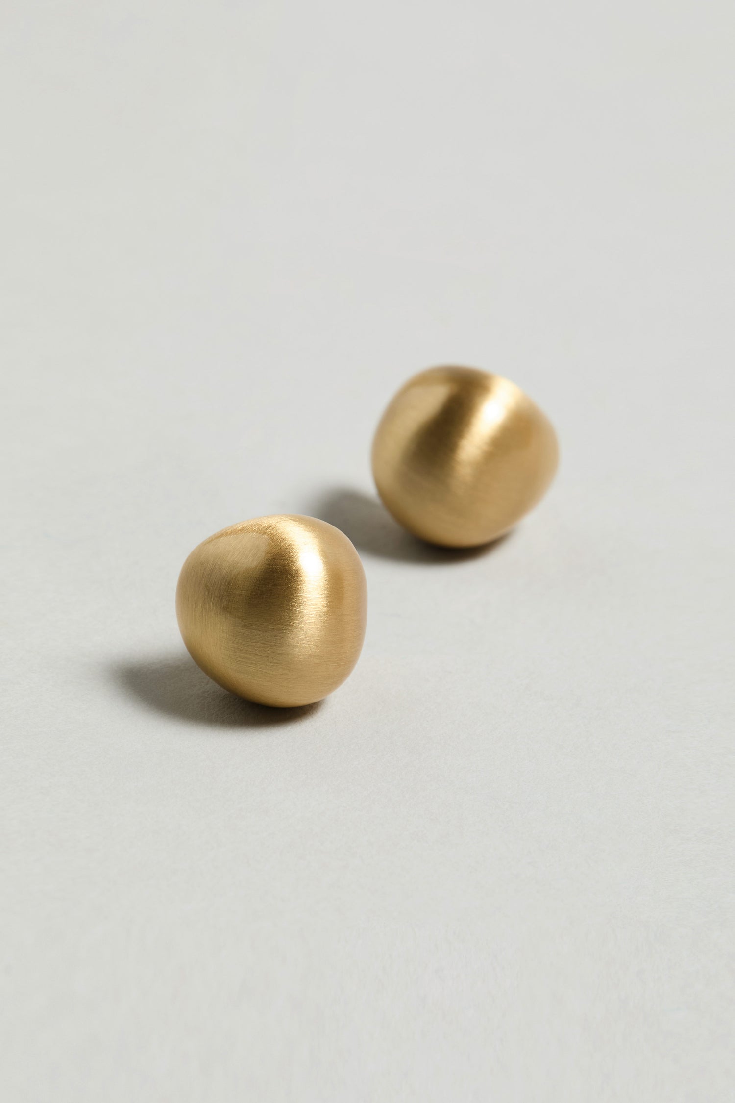 Two Gold Plated Mini Pebble Earrings with a small, rounded shape and matte finish rest on a plain light gray surface.