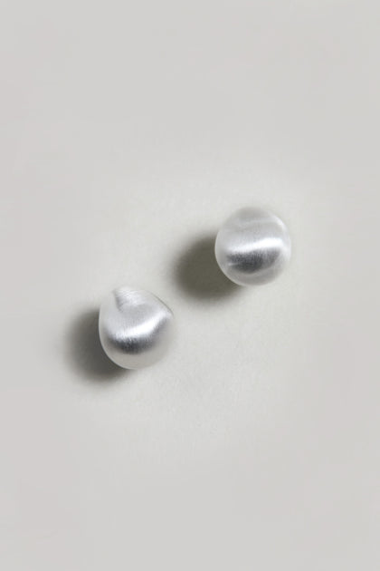 The Mini Silver Pebble Earrings, featuring a round, irregular organic shape and a silver-tone finish, are displayed on a plain light-colored surface.