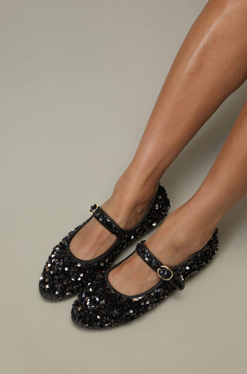 Someone is shown from the knees down wearing Sequin Mary Jane Velvet Shoes—black, handmade in Spain with sequin detail, a strap, and buckle—set against a neutral background.