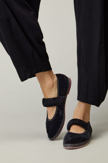 Wearing Mafalda Black Velvet Shoes—handmade in Spain with elastic straps and black velvet, embodying slow fashion—paired with black pants, one foot slightly raised.