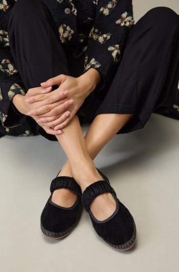 Person sits cross-legged, wearing black pants and Mafalda Black Velvet Shoes—Mary Jane style flats with elastic straps, handmade in Spain—with hands resting on their knees.