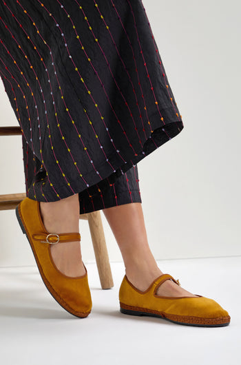 A person wearing Mustard Mary Jane Velvet Shoes, crafted sustainably, and black pants with colorful vertical stitching sits on a wooden chair.