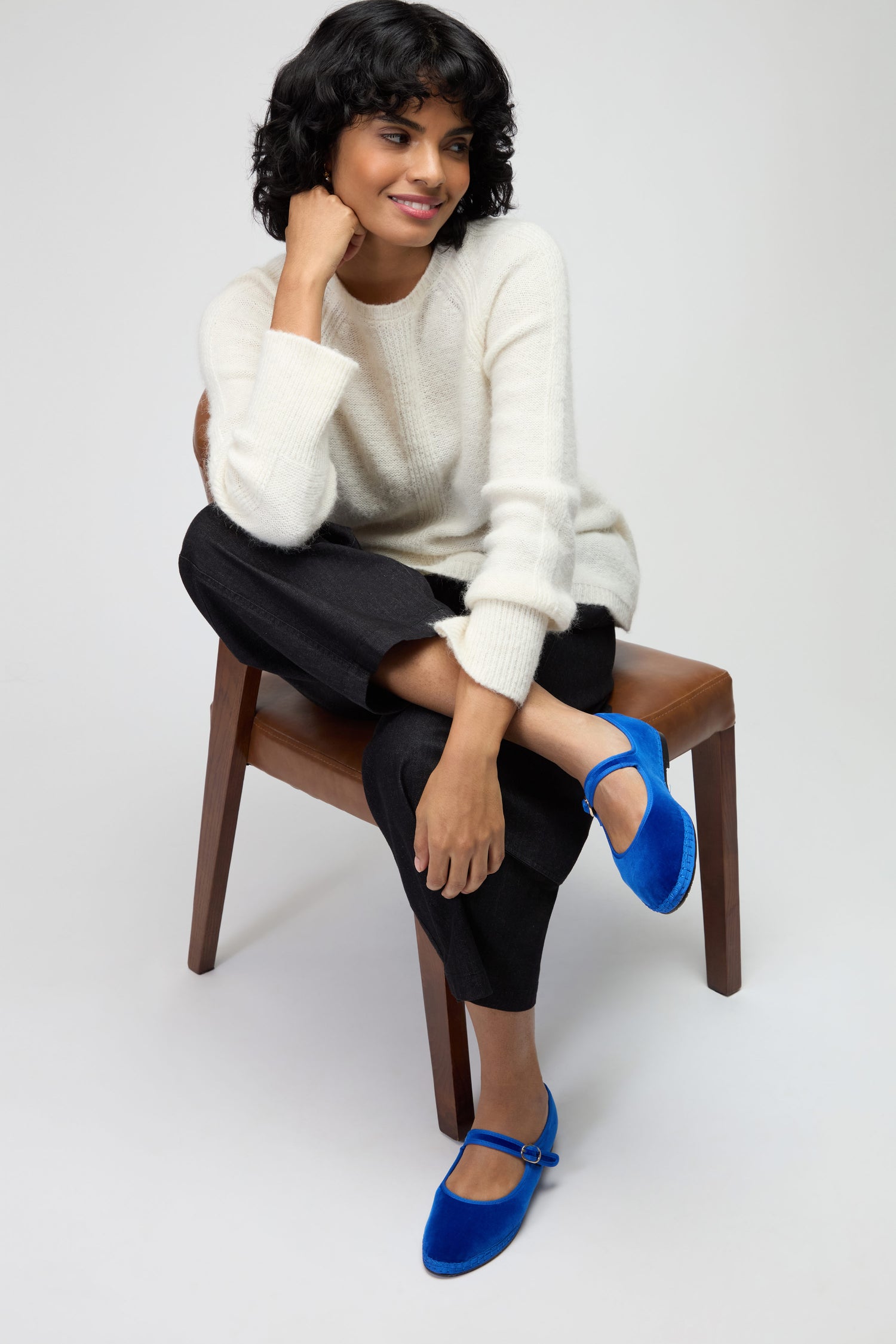 A woman with short curly hair sits on a chair, wearing a white sweater, black pants, and Mary Jane Velvet Shoes—adding a lively touch to her sustainable fashion look.