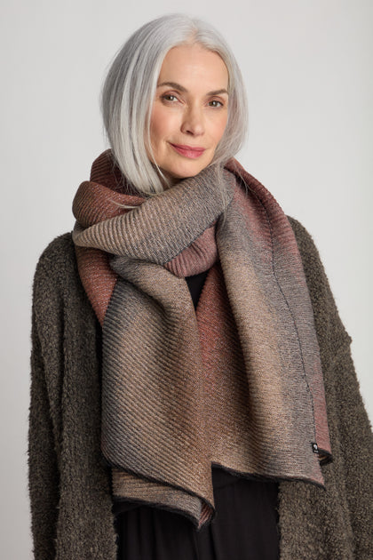 A woman with gray hair wears a large beige and brown Stardust Scarf from Fraas, wrapped around her neck, along with a dark, textured sweater. She is smiling slightly and looking towards the camera.