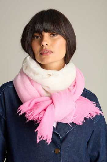 A person with dark, shoulder-length hair is shown wearing a denim shirt complemented by the Ombre Wool Scarf, featuring shades of white and pink. They have a neutral expression and are facing forward against a plain background. This versatile Ombre Wool Scarf adds a touch of elegance to their outfit.