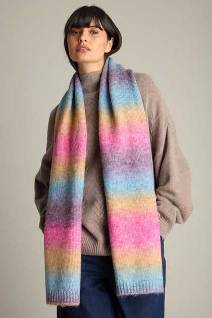 A person in a brown sweater and dark pants is wearing the Rainbow Stripe Knit Scarf, a cozy accessory with horizontal stripes in pastel shades.