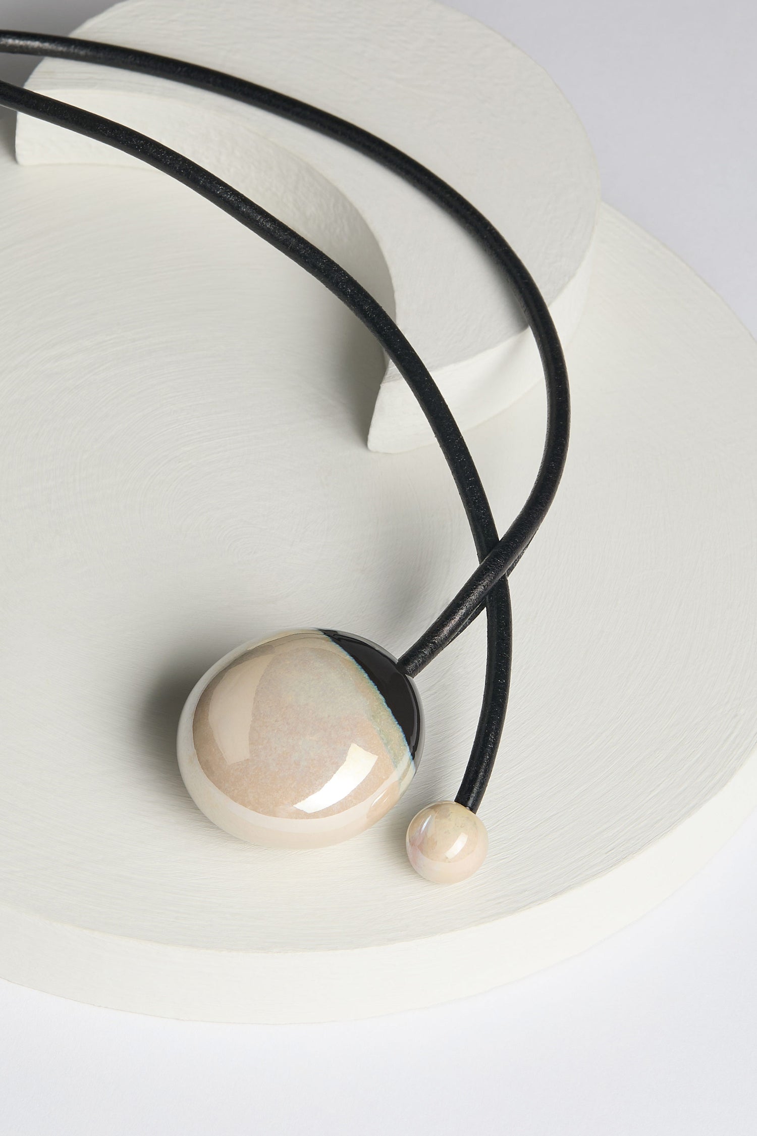 The Handmade Ceramic Pearl Bead Necklace boasts a large beige spherical pendant complemented by a smaller sphere, both elegantly strung on a black cord, exemplifying artisanal craftsmanship against the backdrop of a white geometric surface.