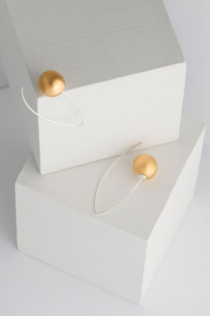 A pair of Handmade Ceramic Mirror Earrings, characterized by their modern, minimalist design with large gold spheres and thin silver wire, featuring 925 sterling silver hooks, beautifully displayed on abstract white blocks.