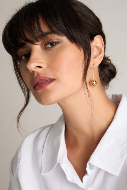 A person with dark hair and bangs, adorned in a white shirt and gold dangling earring, gazes at the camera, embodying modern design with their Handmade Ceramic Mirror Earrings.