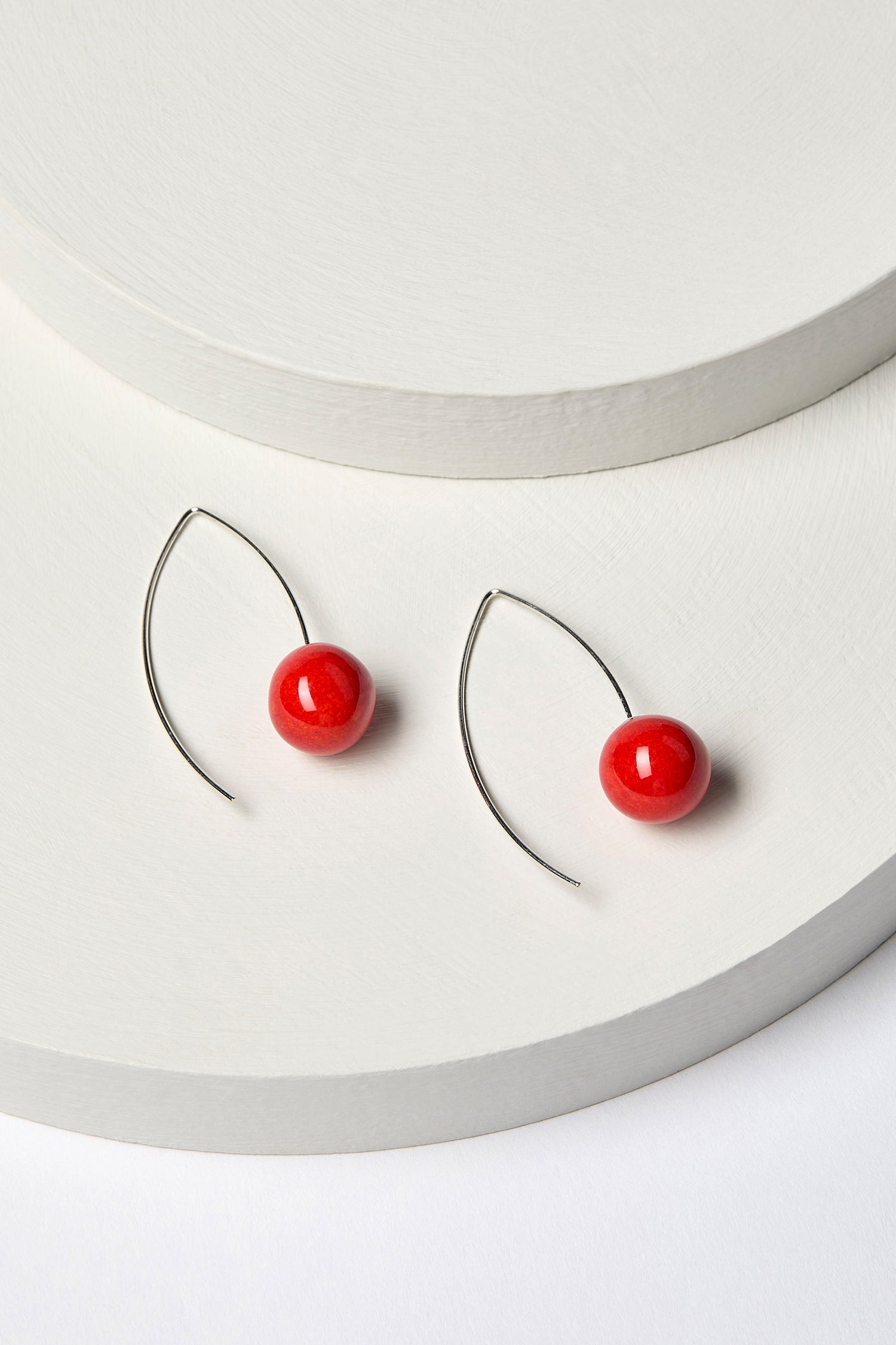 A pair of handmade ceramic bead earrings featuring spherical red beads, crafted with 925 sterling silver wires, elegantly displayed on a curved white surface.