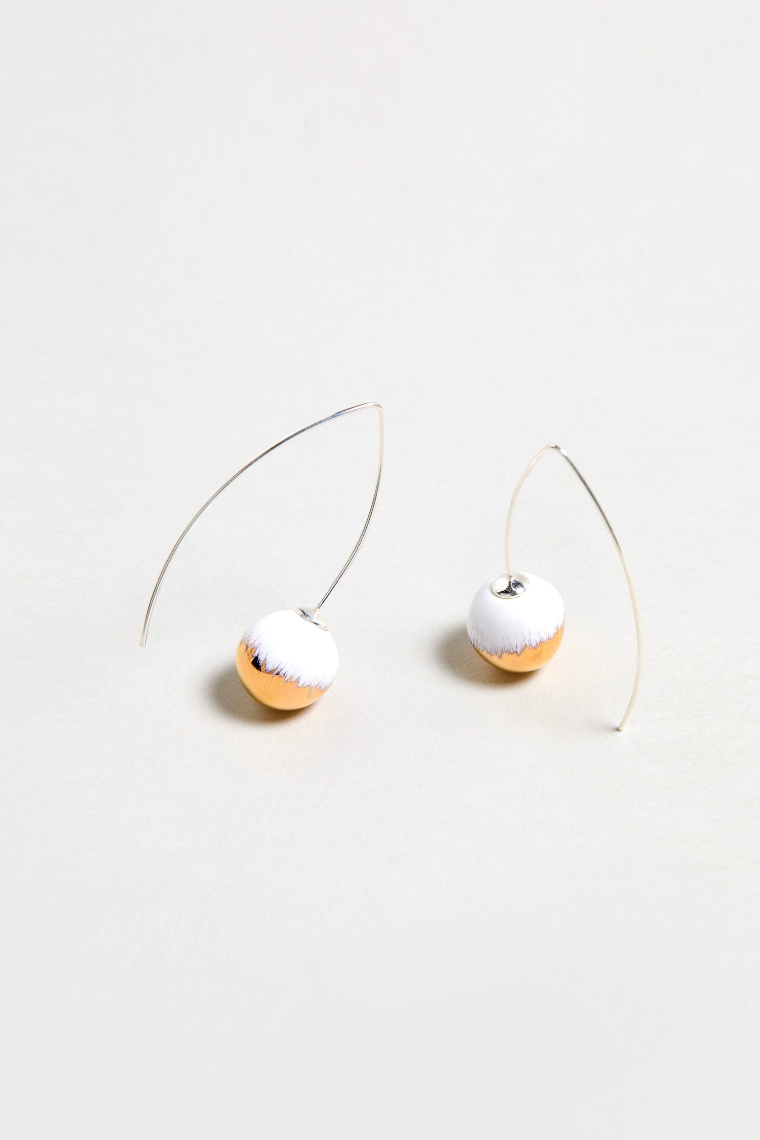 Handmade Gold Brushstroke Ceramic Bead Earrings feature curved silver wires and round, half-white, half-gold ceramic beads. Inspired by gold brushstroke earrings and photographed on a plain light background.
