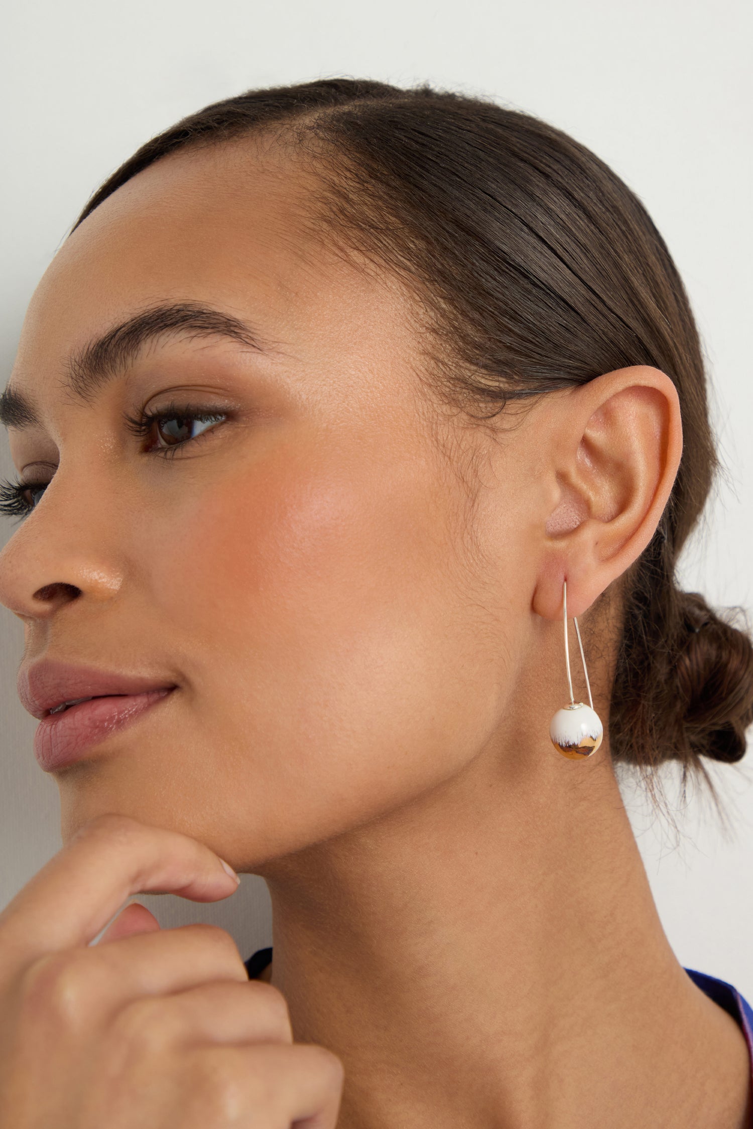A woman with smooth skin and a low brown bun wears Handmade Gold Brushstroke Ceramic Bead Earrings, crafted in France, gazing to the side with her hand near her chin.