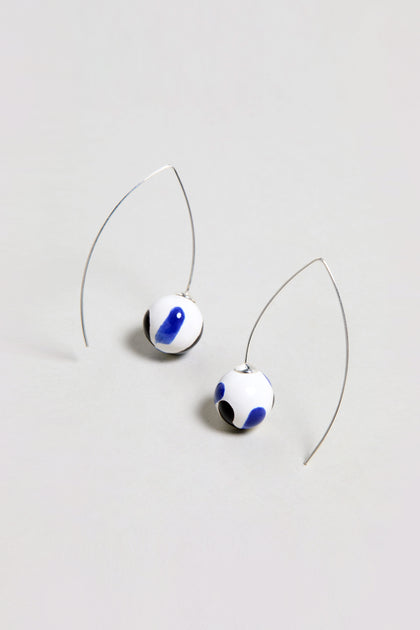 Handmade Painted Ceramic Bead Earrings feature white ceramic spheres with blue and black abstract spots, each elegantly suspended from a curved silver wire.