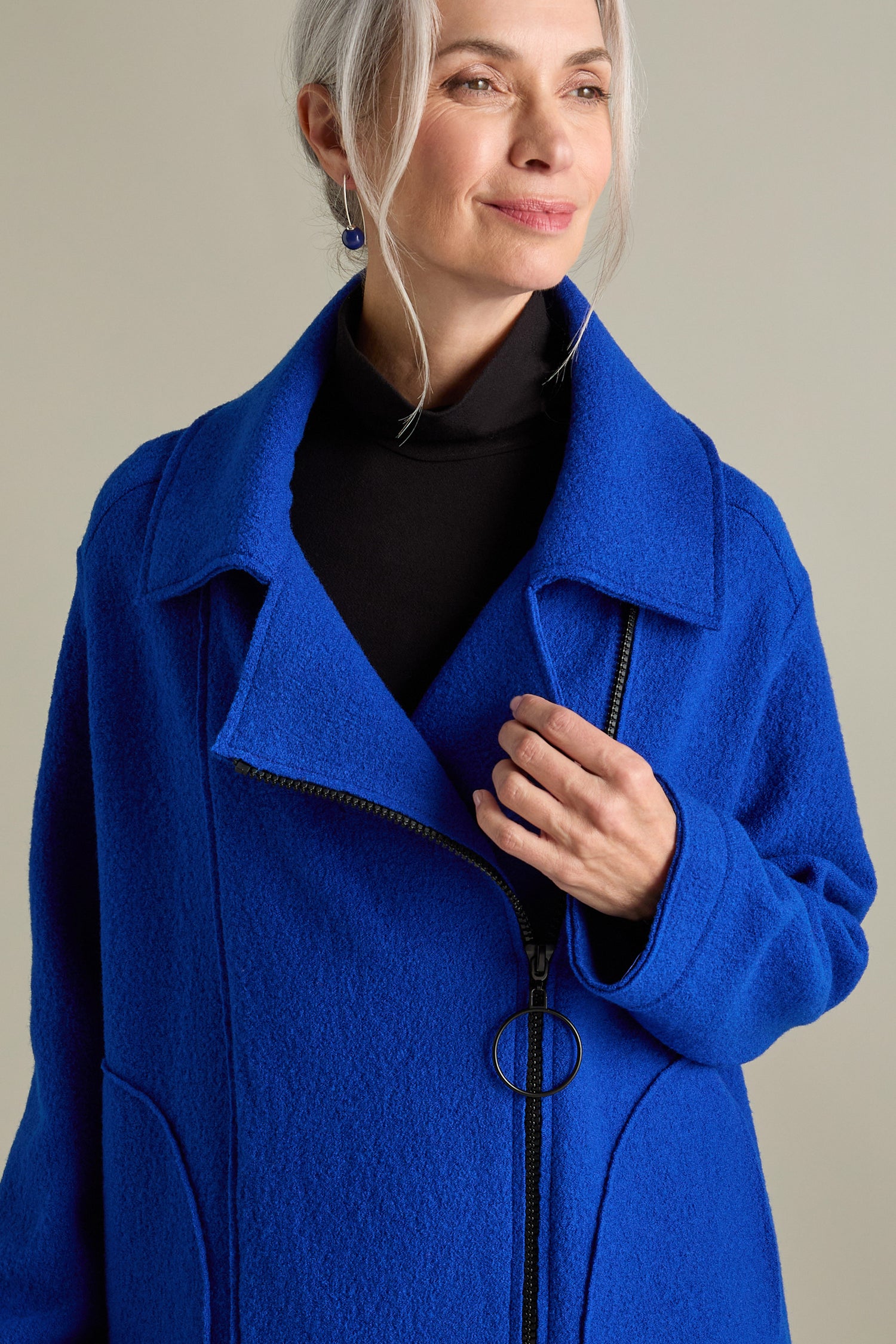 A woman with silver hair wearing a royal blue Boiled Wool Zip Boxy Jacket and a black turtleneck, looking slightly to the side with a serene expression.