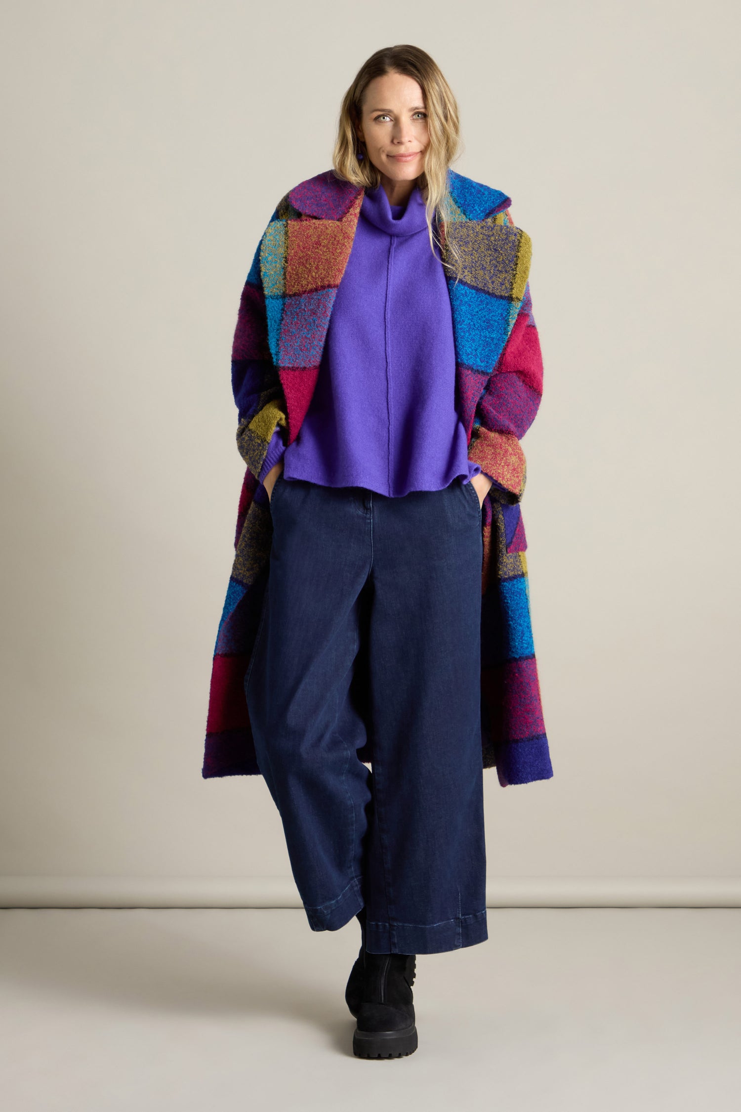 Person wearing a vibrant statement piece: a Check Wool Coat made of a colorful boucle wool blend over a purple sweater and wide-leg jeans, standing against a plain background.