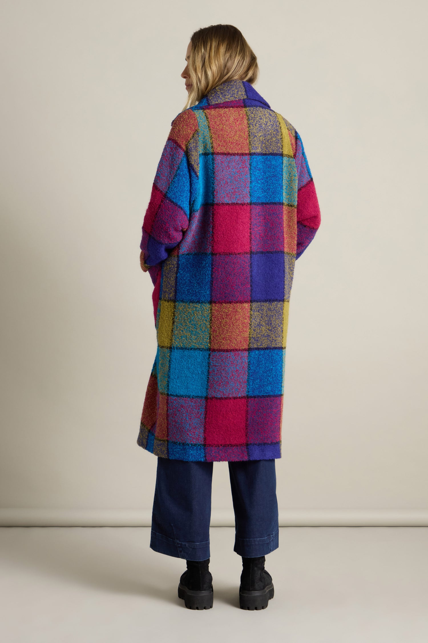 A person stands facing away, wearing a vibrant statement piece—the Check Wool Coat, featuring a long, colorful checkered pattern in shades of pink, blue, purple, and yellow. This eye-catching coat pairs perfectly with navy pants and black boots.