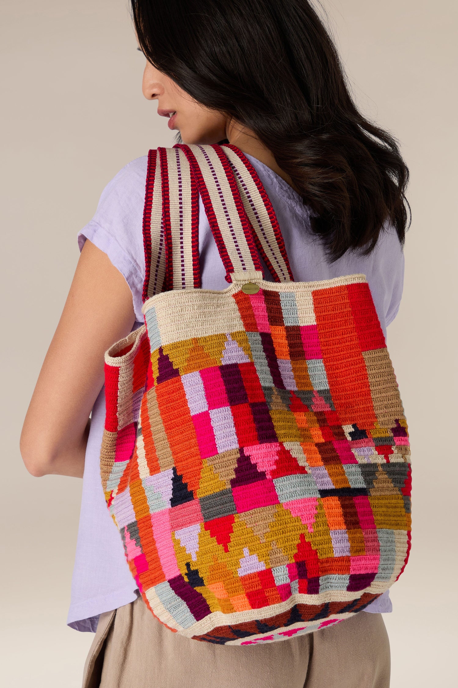 A person with shoulder-length dark hair, wearing a light purple shirt and beige pants, carrying a Handcrafted Cala Tote Bag over their shoulder that showcases traditional techniques and intricate geometric patterns.