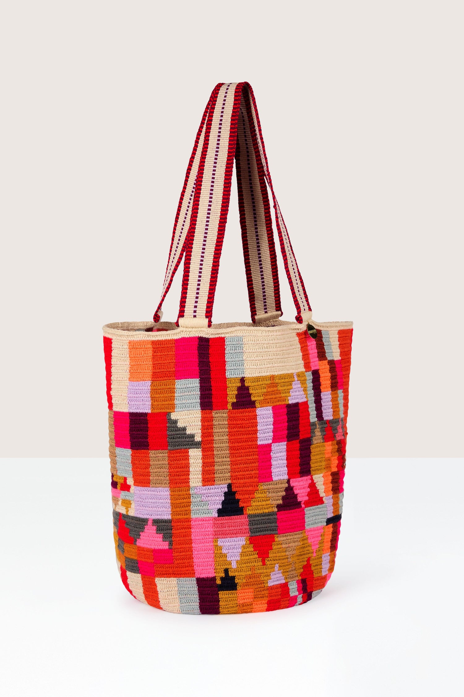 A Handcrafted Cala Tote Bag with a colorful, geometric pattern in shades of red, pink, orange, and beige, featuring striped handles and created using traditional techniques.