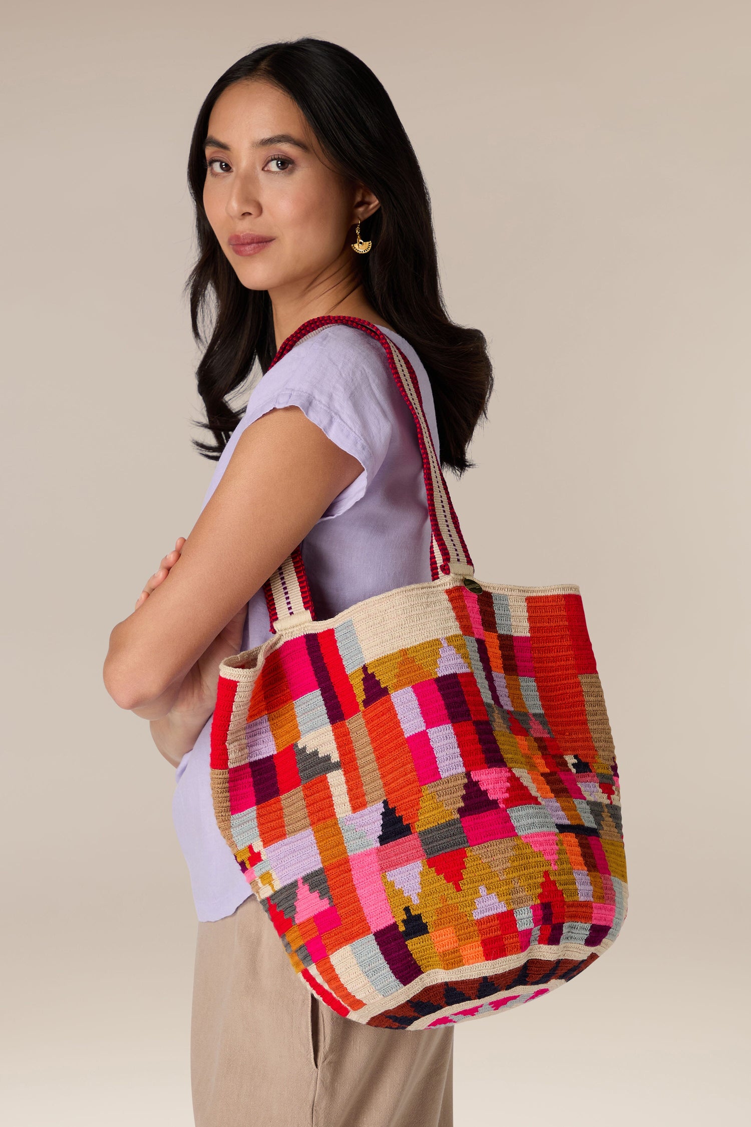 Woman with long dark hair wearing a light purple shirt, beige pants, and gold earrings, holding a colorful Handcrafted Cala Tote Bag with geometric patterns.