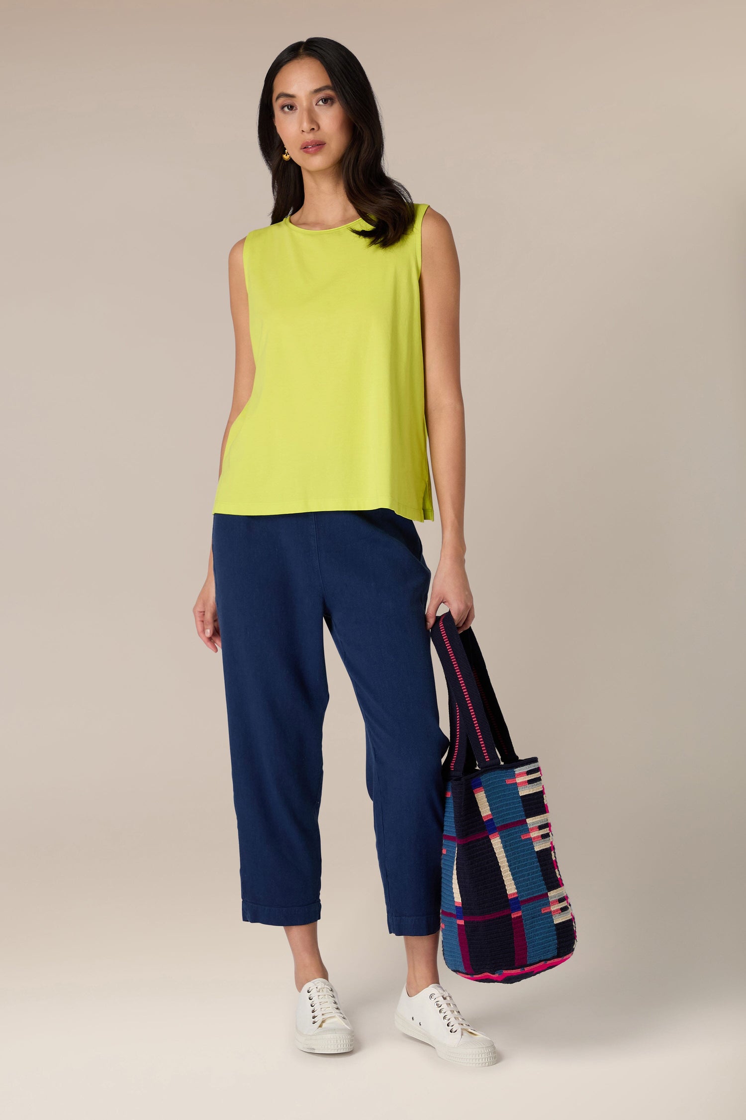 A woman with long black hair is wearing a yellow sleeveless top, blue pants, and white sneakers. She is holding a Handcrafted Cala Tote Bag adorned with geometric patterns, crafted using traditional techniques, and standing against a plain background.