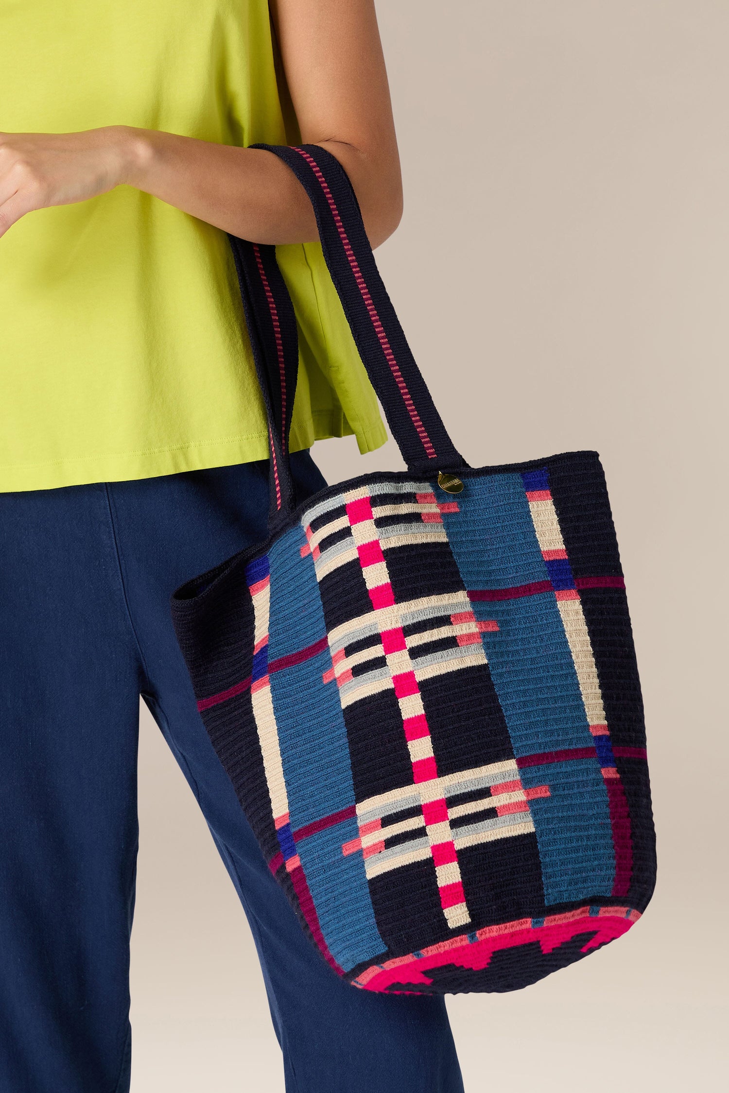 Person wearing a yellow top and blue pants, holding the Handcrafted Cala Tote Bag featuring a blue, pink, and white geometric pattern created by skilled artisans.