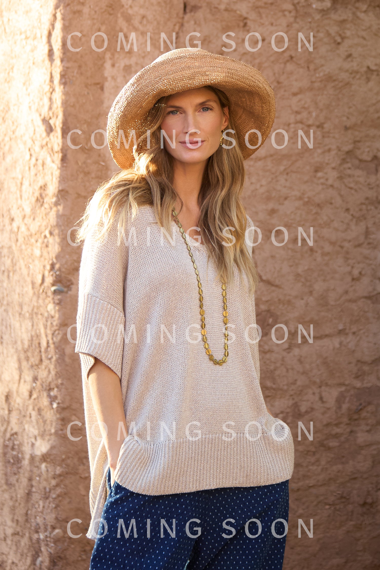 A woman in a wide-brimmed straw hat, V Neck Relaxed Knit Top, and blue pants stands by a textured brown wall with "COMING SOON" and code GOK2964-VIY displayed.
