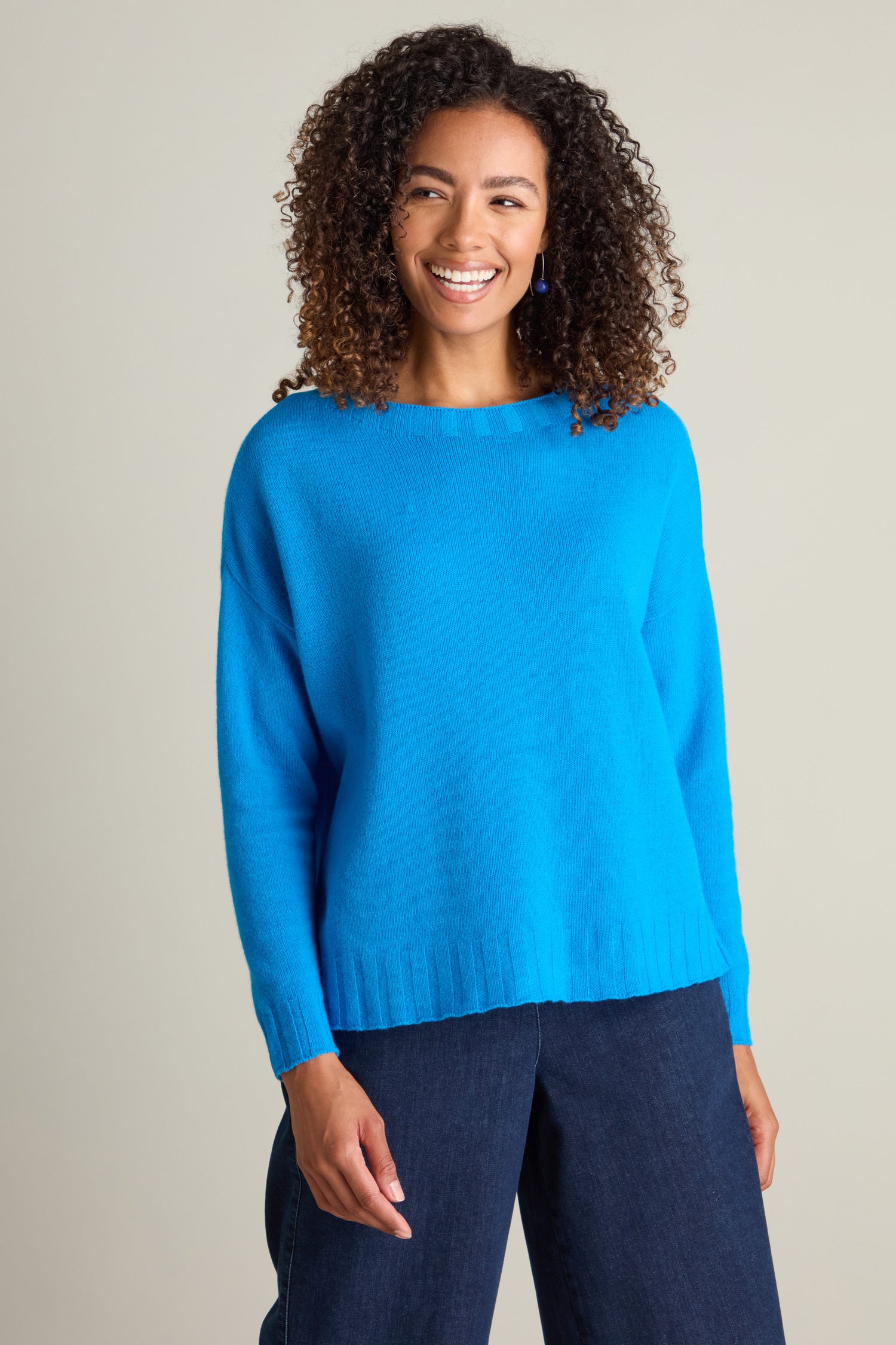 A person with curly hair smiles while wearing a bright blue Cashmere Blend Crop Jumper and dark blue pants.