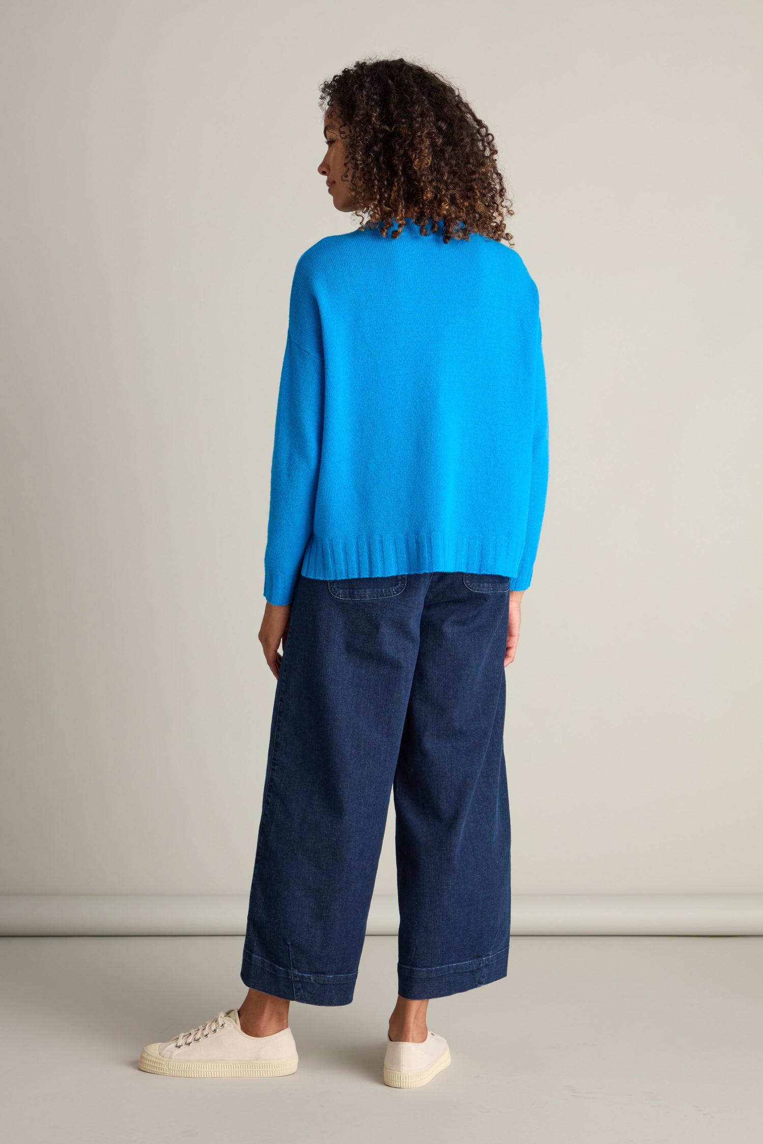 Person with curly hair wearing a bright blue Cashmere Blend Crop Jumper, dark blue wide-leg pants, and white sneakers, standing and facing away from the camera against a plain background.