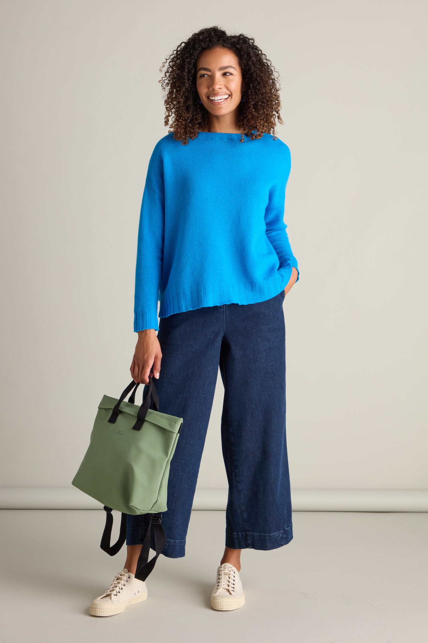 A person stands smiling, wearing a blue Cashmere Blend Crop Jumper with its cropped silhouette, dark wide-leg pants, and beige sneakers. They hold a green bag in their left hand and have their right hand in their pocket against a plain background.