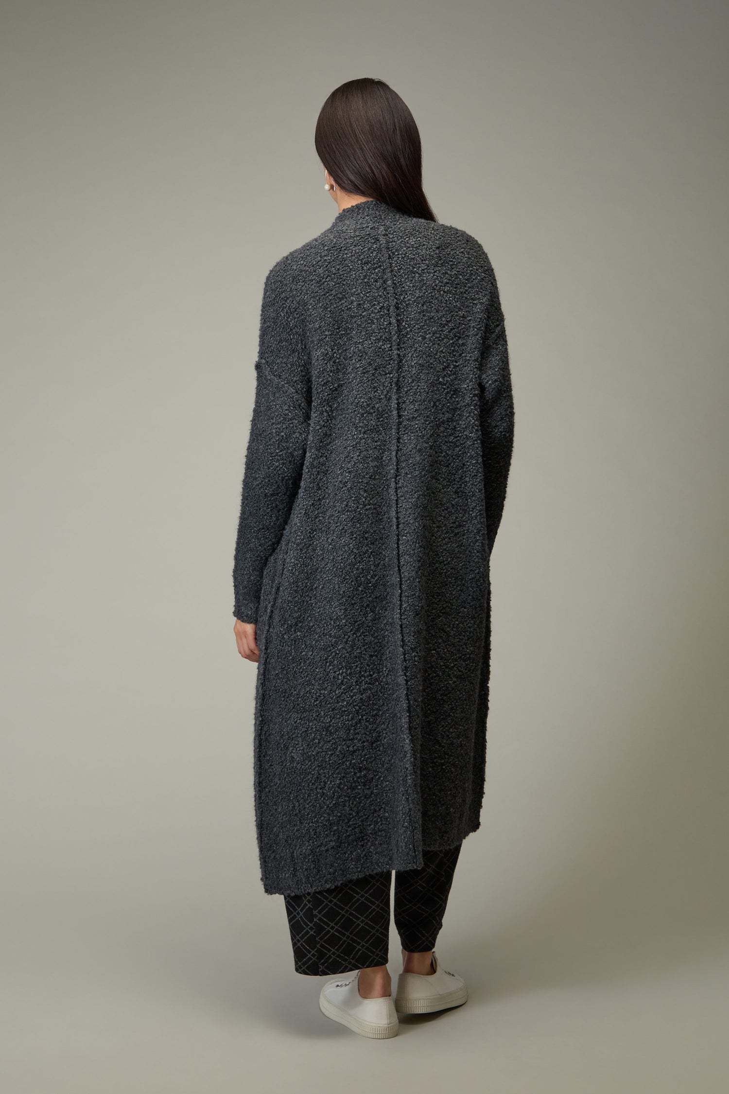 A person stands with their back to the camera in a Charcoal Boucle Knit Long Cardi, black pants, and white shoes against a plain backdrop—an ideal choice for updating your autumn wardrobe.