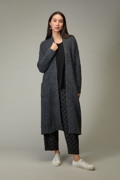 A woman stands against a plain backdrop wearing the Charcoal Boucle Knit Long Cardi, a black top, patterned black pants, and white sneakers—making this cardi a stylish essential for any autumn wardrobe.