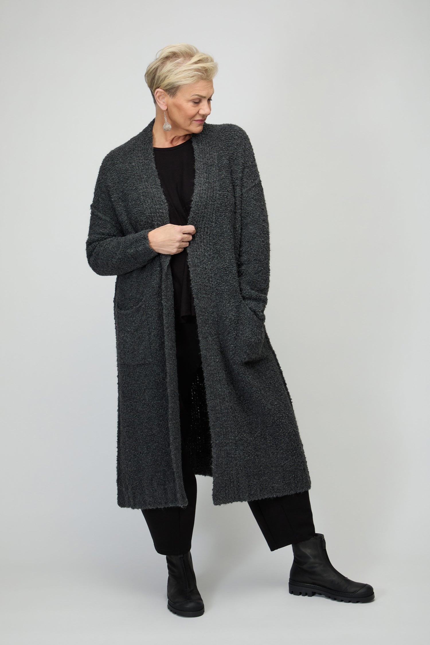 A person with short blond hair wears the Charcoal Boucle Knit Long Cardi with black pants and boots, standing against a plain light gray background—an ideal look for effortless autumn layering.