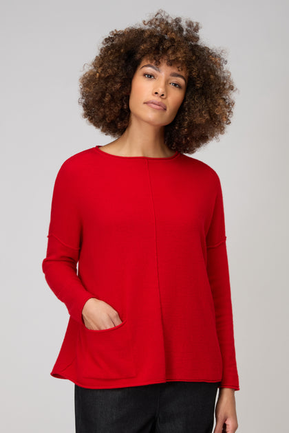 A curly-haired person models the Merino Pocket Jumper in red, with a front pocket and long sleeves, standing against a plain light background.