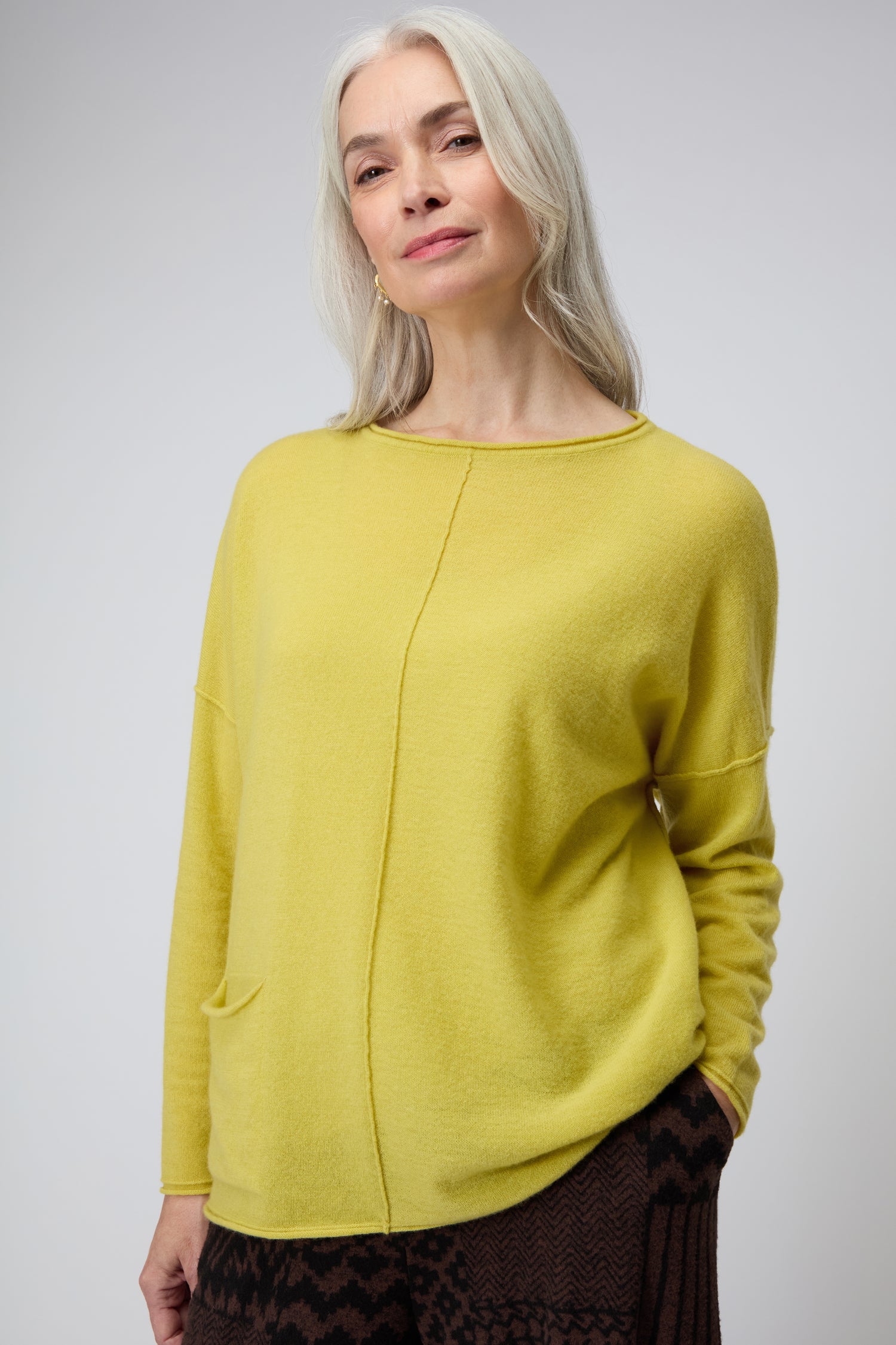 A woman with long gray hair wears a yellow Merino Pocket Jumper and patterned brown pants, standing against a plain light background.