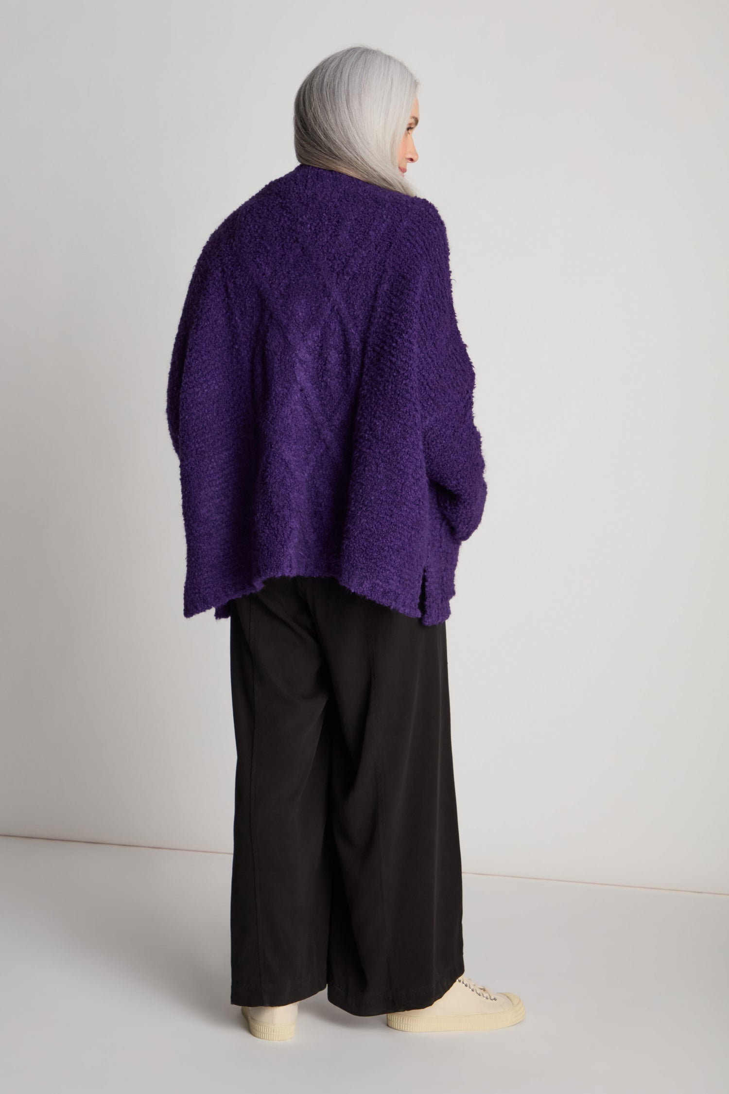 A person with long gray hair is standing in profile, wearing a loose-fitting purple Bouclé Knit Cable Trim Cardi, black wide-leg pants, and off-white shoes.