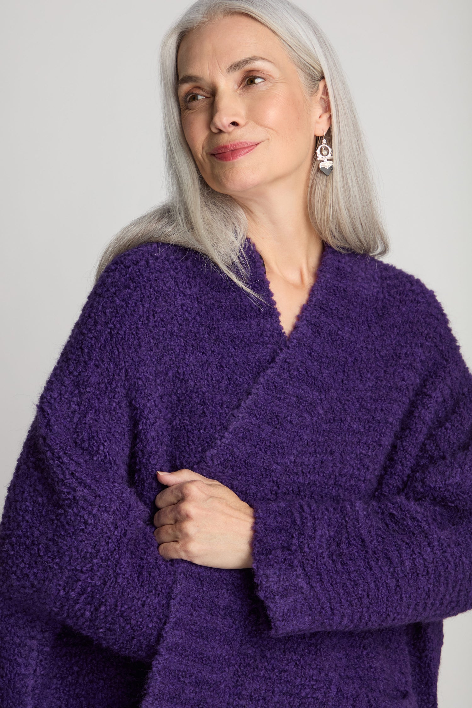 A woman with long gray hair, wearing a Boucle Knit Cable Trim Cardi adorned with silver earrings, looks to the side with a slight smile.